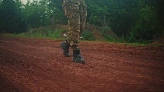 Camouflage pants and rugged boots in walking motion along red dirt path beside dense green forest, low angle ground view showing earthy texture, outdoor terrain, and focused movement