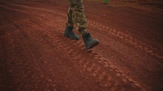 Camouflage trousers and hiking boots stepping along reddish dirt road in rugged outdoor terrain with low viewpoint, earthy colors, soft natural light, and active walking motion