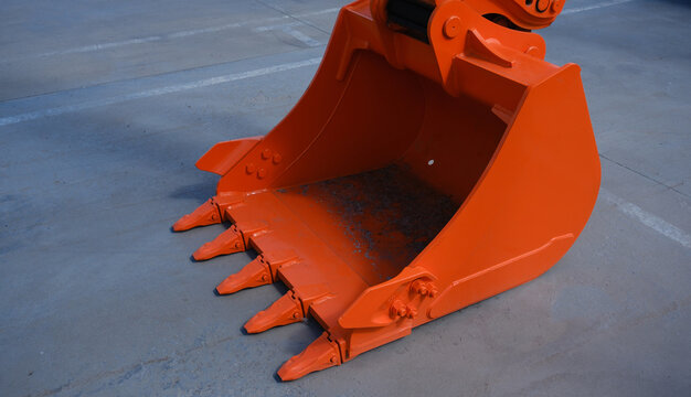 The teeth are new. The excavator bucket is used to move soil using a mechanical arm. The equipment is painted orange on a summer day.