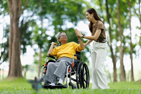Asian woman caregiver high-fiving elderly patient in wheelchair, showing support, care, and achievement during an outdoor park activity, promoting independence and happiness