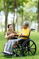 Obraz premium Young caregiver providing comforting support to an elderly asian woman using a wheelchair while enjoying a peaceful day outdoors in a green park, focusing on assistance and senior care