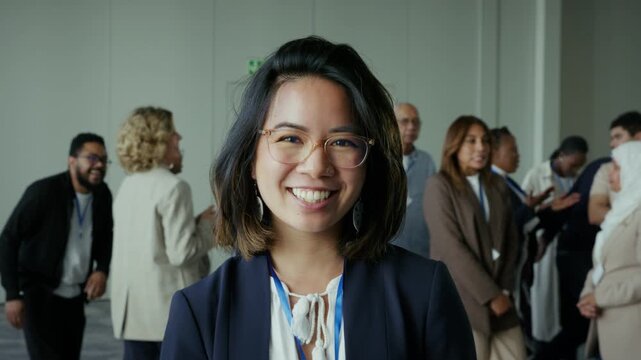 A confident young Asian woman smiles brightly at a busy professional business conference. Diverse attendees are seen networking and engaging in the background.