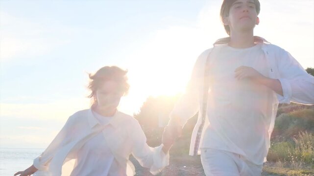 Happy young boy and girl in love running hand in hand along seashore at sunset. Teenage brother and sister in white clothes running away from the sun in slow motion. Carefree and beloved young people