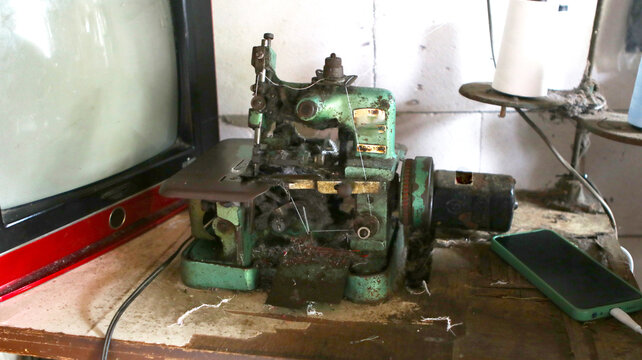 Old vintage green overlock sewing machine on a dusty workshop table. The machine sits on a dusty wooden workbench next to a smartphone with a green case and an old CRT television set on the left