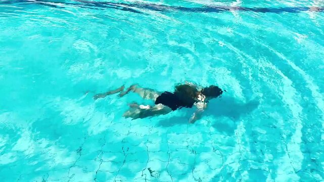 Teenage girl in a black swimsuit swimming underwater in a clear blue pool. Represents summer fun, youth, fitness, freedom, and aquatic activity.
