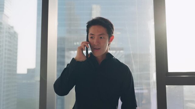 Man in Office Making a Call Receiving Good News: A focused man stands near a large window in a modern office, engaged in a phone conversation with a city skyline visible in the background.
