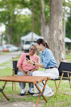 Young caregiver explaining mobile technology to senior woman, a happy retired grandmother learning while using a smartphone with assistance outdoors in a park