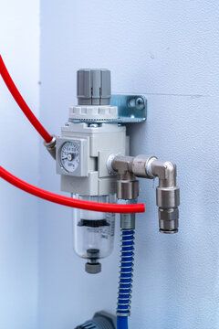 Close-up of pneumatic air pressure regulator with gauge, metal fittings, and red tubing mounted on a wall.