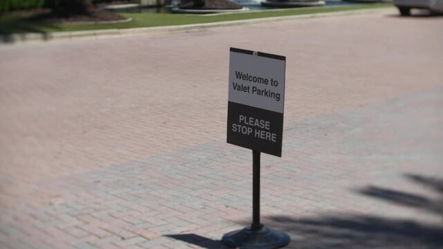 Person carrying shopping bags beside a valet parking sign near a parked SUV on a sunny day. Luxury retail destination entrance with customer arriving at curbside parking drop-off area