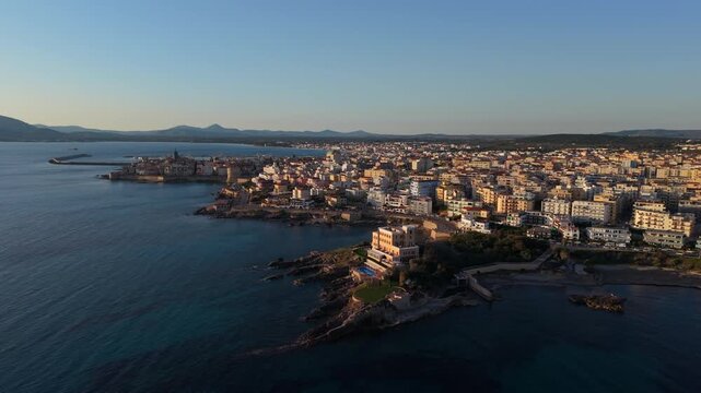Mediterranean city blocks line the rocky waterfront as the camera glides along the Alghero shoreline in warm evening light.