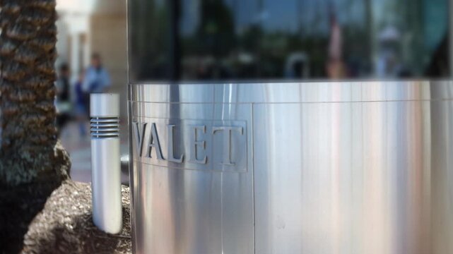 Valet sign engraved on a polished metal structure outside a commercial building entrance. Close-up detail of parking service signage with shallow depth of field and blurred pedestrians