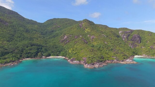 Drone footage revealing a hidden white sand beach nestled between lush tropical green mountains and crystal-clear turquoise waters in anse major and anse du Riz Mahe, Seychelles.