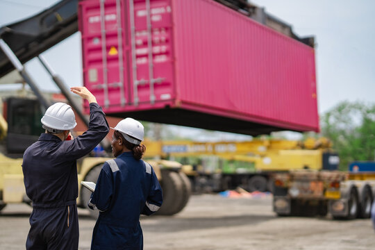 Logistics workers guide container handling operation while watching heavy equipment move freight. Team communication helps keep loading process organized and on schedule.