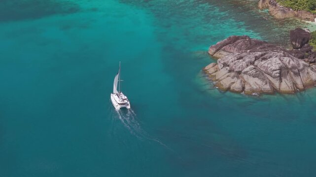 High-angle drone shot of a luxury catamaran cruising over a clear turquoise coral reef past granite boulders in anse du Riz, Mahe, Seychelles.