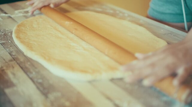 Close-up of baker hands using a wooden rolling pin to roll out and flatten fresh yeast dough. Preparing homemade sweet pastry or cinnamon rolls on a floured table, 4k.