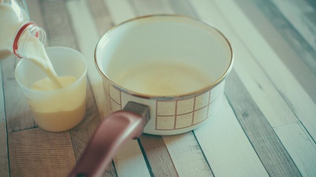 Close-up of pouring fresh milk from a bottle into a plastic measuring cup on a wooden table. Measuring liquid ingredients next to a small pot for a sweet pastry recipe, 4k.