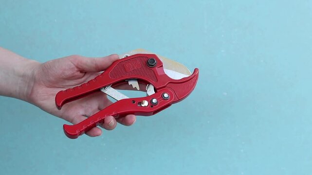 Hand holds red pipe cutter for plastic pipes on blue background. Professional hand tool. Hand squeezes red scissors for cutting PVC and polypropylene pipes. Master holds tool for installing water supp