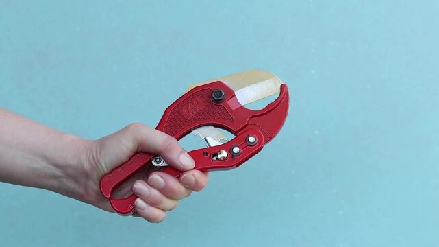 Hand holds red pipe cutter for plastic pipes on blue background. Professional hand tool. Hand squeezes red scissors for cutting PVC and polypropylene pipes. Master holds tool for installing water supp