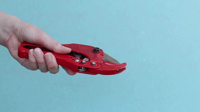 Hand holds red pipe cutter for plastic pipes on blue background. Professional hand tool. Hand squeezes red scissors for cutting PVC and polypropylene pipes. Master holds tool for installing water supp