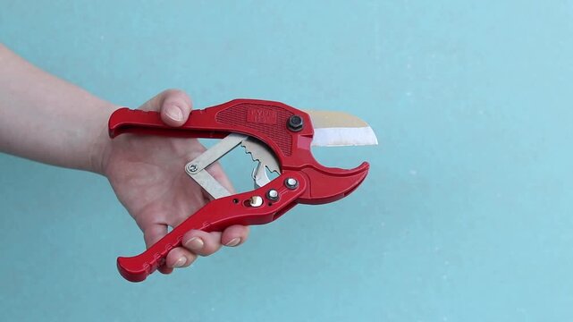 Hand holds red pipe cutter for plastic pipes on blue background. Professional hand tool. Hand squeezes red scissors for cutting PVC and polypropylene pipes. Master holds tool for installing water supp