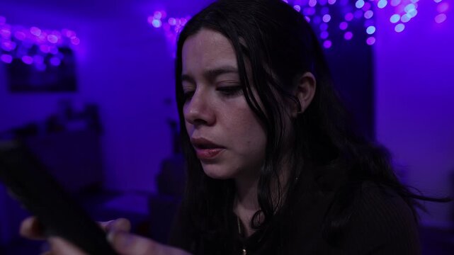 A young woman intently uses her smartphone in a moody, dimly lit room, illuminated by glowing purple LED string lights. The close-up shot reveals a pensive expression and low-key, cinematic lighting.