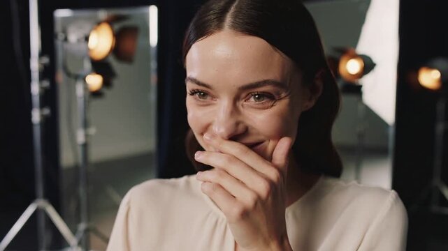 Smiling woman wearing blouse, covering mouth, laughing while camera rolling in studio, lights