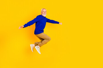Fototapeta premium Happy senior man jumping in blue sweater against bright yellow background, energetic lifestyle portrait of a joyful retired active adult