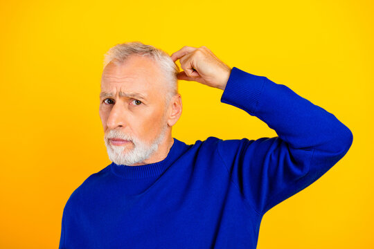 Confused senior man scratching head in blue sweater on yellow background expressing doubt and puzzlement