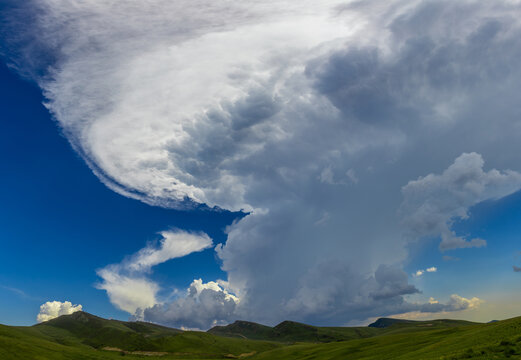 A large cumulonimbus cloud with a distinct anvil shape towering over the green, rolling hills. A massive cumulonimbus incus cloud, commonly known as an anvil cloud or thunderhead