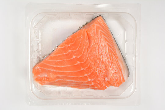 Top view of a fresh raw salmon fillet with skin packed inside a transparent plastic grocery container on a white background