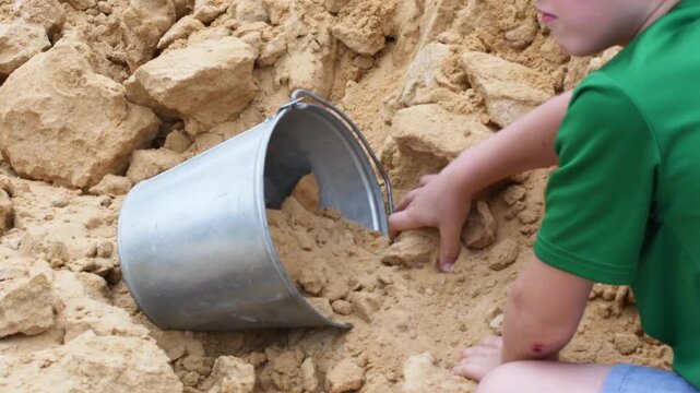 Young boy in a green shirt kneeling and playing with his hands in a sandbox, carefully scooping up handfuls of sand and putting them inside a small galvanized metal pail for fun