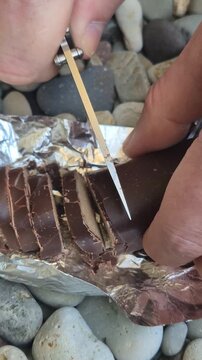 Chocolate knife cutting Close up of hands slicing a chocolate bar with a folding knife on a foil wrapper over pebbles