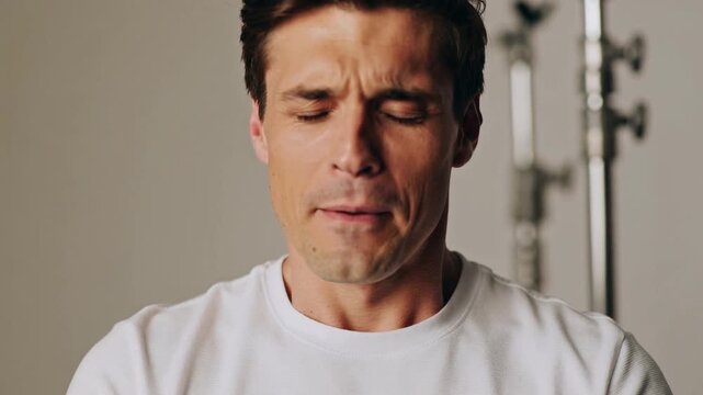 Smiling man in white tee responding to off-camera cue, closing eyes, pursing lips in studio stands
