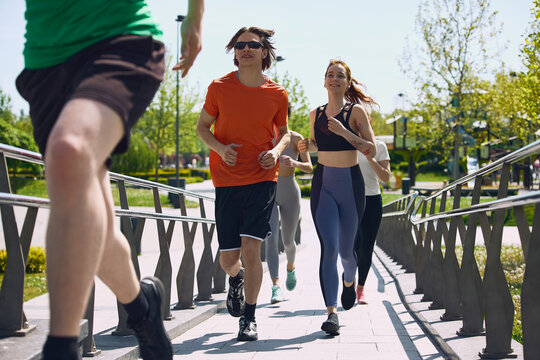 Young adults running across pedestrian bridge during outdoor cardio training session. Concept of endurance training, healthy habits, active lifestyle, fitness community, teamwork and urban recreation.