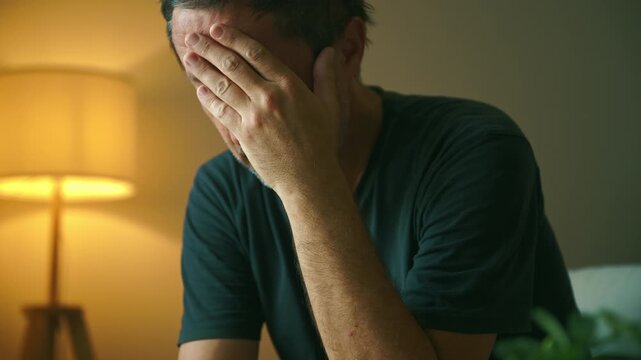 A weary man sits in a dimly lit living room, covering his face with his hand as a warm lamp glows behind him, conveying stress, exhaustion and emotional struggle during late-night hours. 4K.