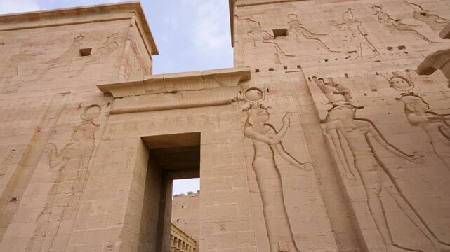 A low angle view of the ancient stone pylon entrance at the Temple of Philae in Aswan, Egypt, featuring intricate hieroglyphic carvings and goddess reliefs.