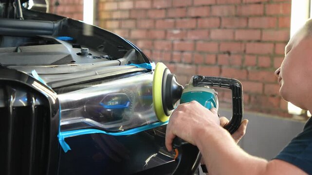 Man polishes the glass of a car headlight with a polishing machine from small scratches, chips. Process polishing of the front car headlights. Hands of male professional worker with orbital polisher. 