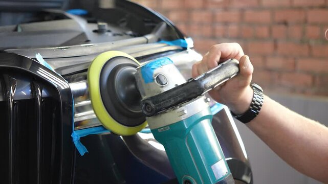 Man polishes the glass of a car headlight with a polishing machine from small scratches, chips. Process polishing of the front car headlights. Hands of male professional worker with orbital polisher. 