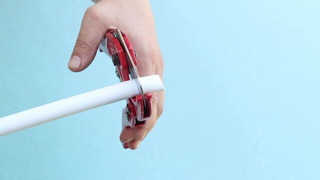 The process of cutting a white plastic pipe with a red pipe cutter. The master uses a manual ratchet pipe cutter to cut a PVC pipe on a blue background. Close-up of a tool in operation