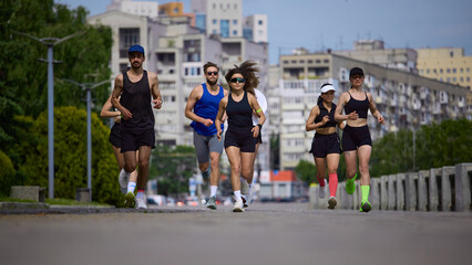 Fototapeta premium Running club jogging toward camera on urban road with city buildings behind. Concept of city fitness, cardio training, endurance, active lifestyle and group motivation.
