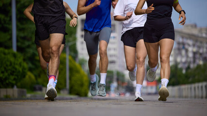 Naklejka premium Cropped legs of running group moving fast on urban road in summer. Concept of cardio workout, running pace, endurance training, sportswear and active lifestyle.