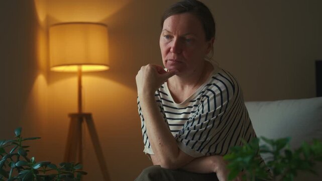 Middle-aged woman with a serious expression deep in thought in a comfortable living room. Contemplation, life choices and mental wellbeing theme. 4K.