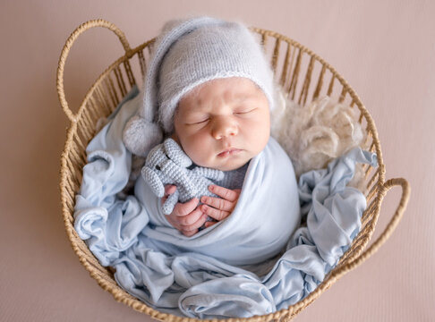 Sleeping newborn rests in cozy basket. Soft blue swaddle, knitted hat, cuddles tiny crochet bunny. Professional newborn photoshoot.