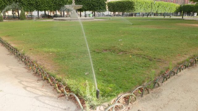 Automated lawn sprinkler watering a green grass lawn in a public park in Paris, with a stone fountain and trees in the background.