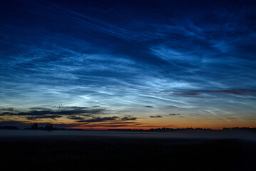 Obraz premium Glowing noctilucent clouds above a misty rural landscape at blue hour, creating a serene and dramatic summer twilight scene.