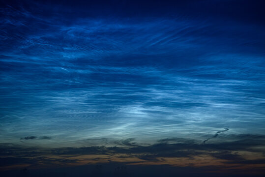 Glowing noctilucent clouds above a misty rural landscape at blue hour, creating a serene and dramatic summer twilight scene.