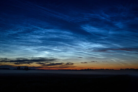 Glowing noctilucent clouds above a misty rural landscape at blue hour, creating a serene and dramatic summer twilight scene.