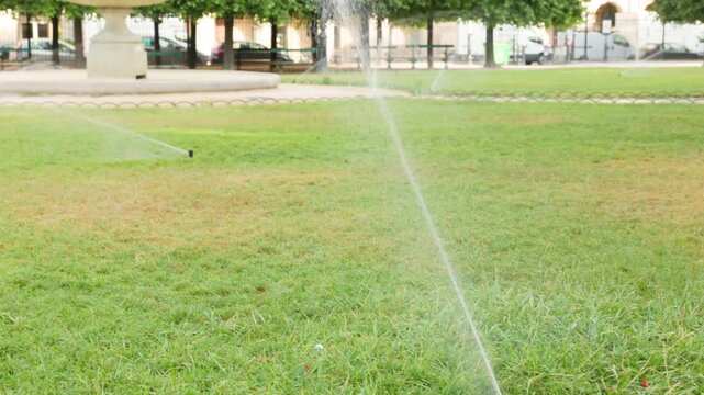 Scenic view of automated lawn sprinklers watering a lush green grass field in a classic Parisian park, featuring a historic tiered stone fountain and manicured green trees in the background.