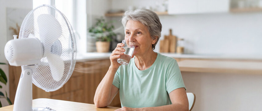 Elderly woman drinks water near a fan for relief from heat, showing a relatable scene of comfort and cooling during hot weather, perfect for lifestyle and health content