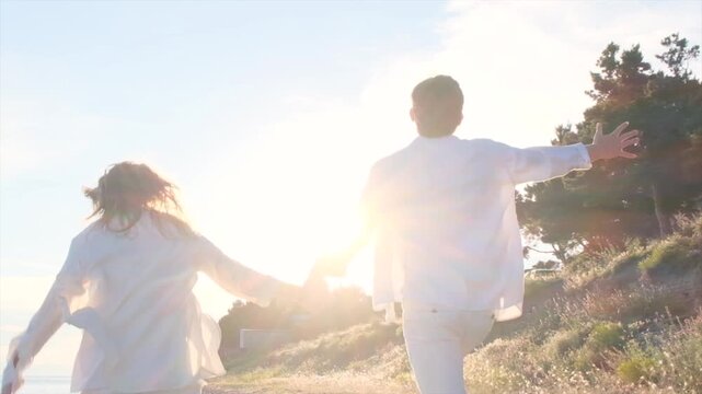 Camera following young boy and girl in white clothes running hand in hand toward sunset in slow motion. Young lovers alone in nature. Concept of first love in youth. Happy brother and sister